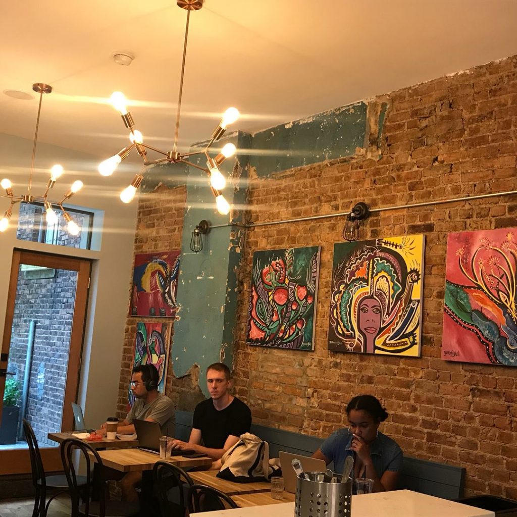 Interior of restaurant with colorful painting hanging from an exposed brick wall and an ornate lamp over the tables at Brown Butter NYC