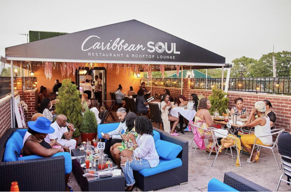 Patrons seated outdoors on chairs covered with blue pillows; black tent with the Caribbean Soul sign covering guests at Caribbean Soul Rooftop Restaurant and Lodge