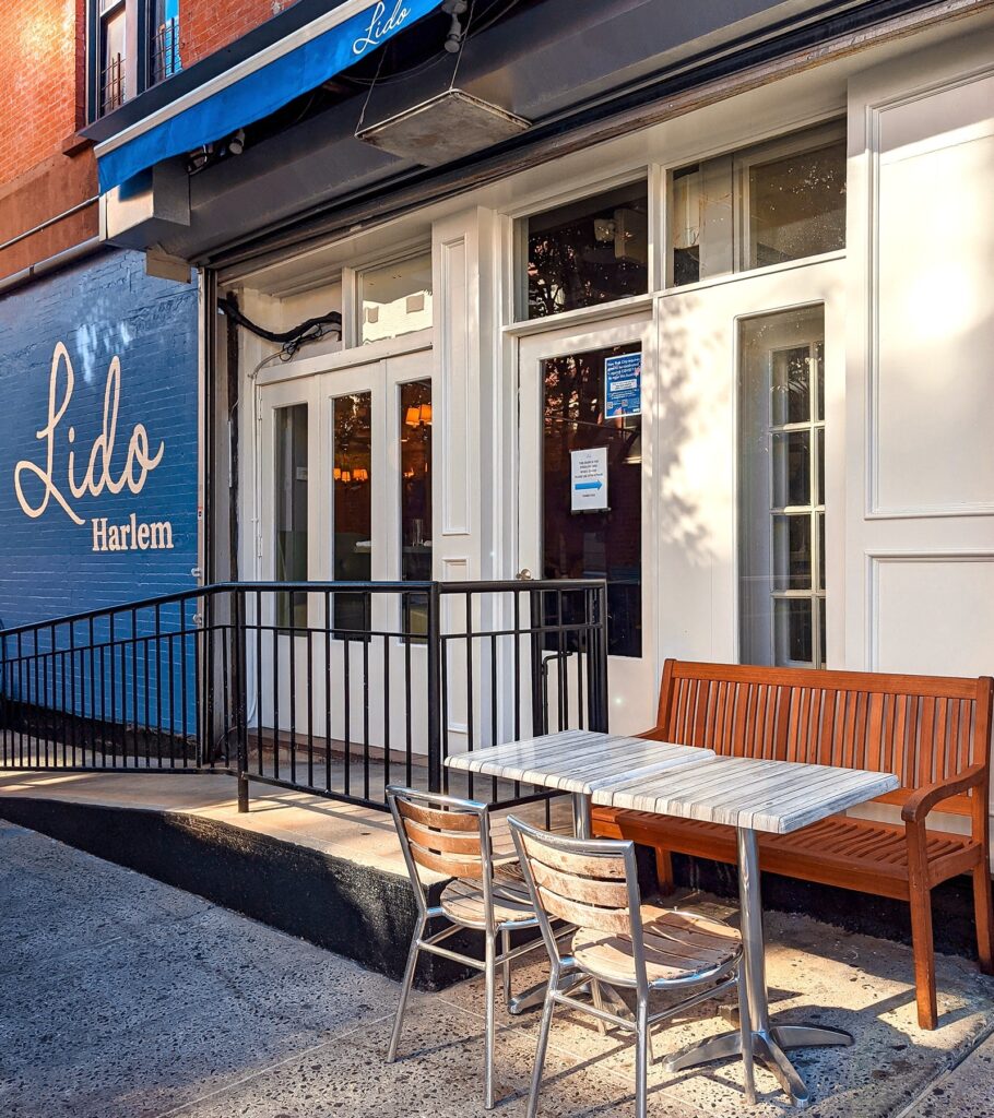 Brown lawn chair and a white table placed outside a restaurant with the Lido signage written with gold letters on a blue background at Lido Harlem Restaurant