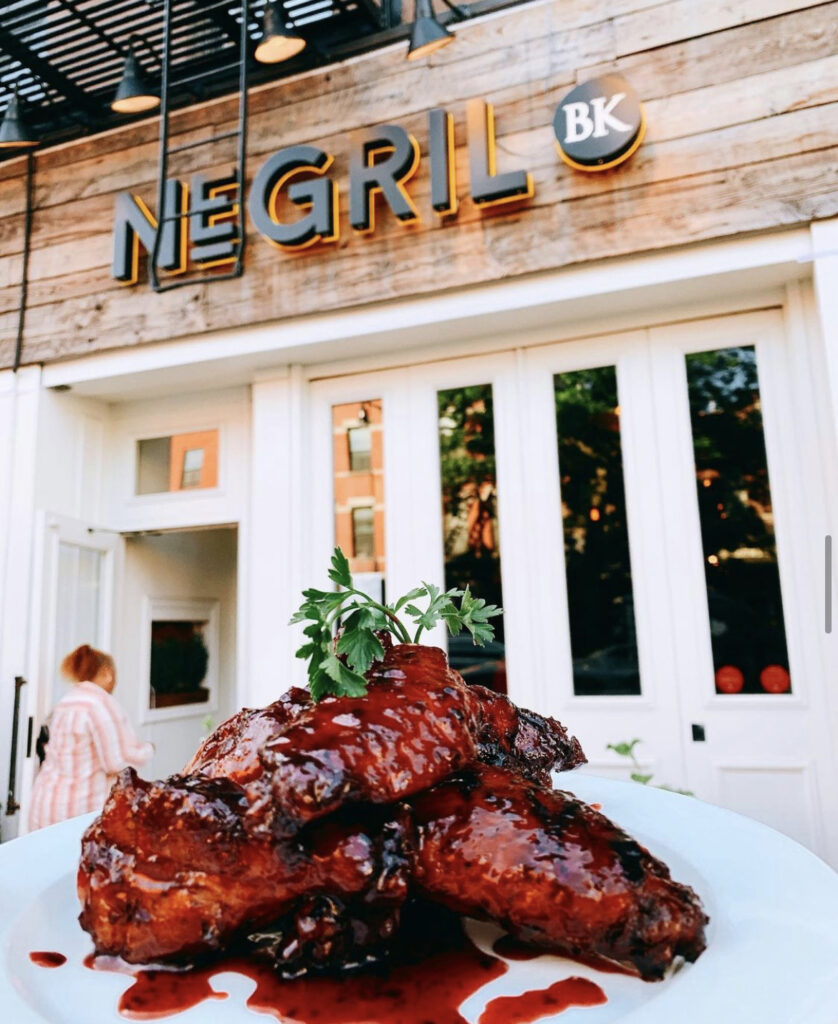 A plate of wings photographed in front of restaurant with white walls and Negril name in black against a wooden background at Negril BK