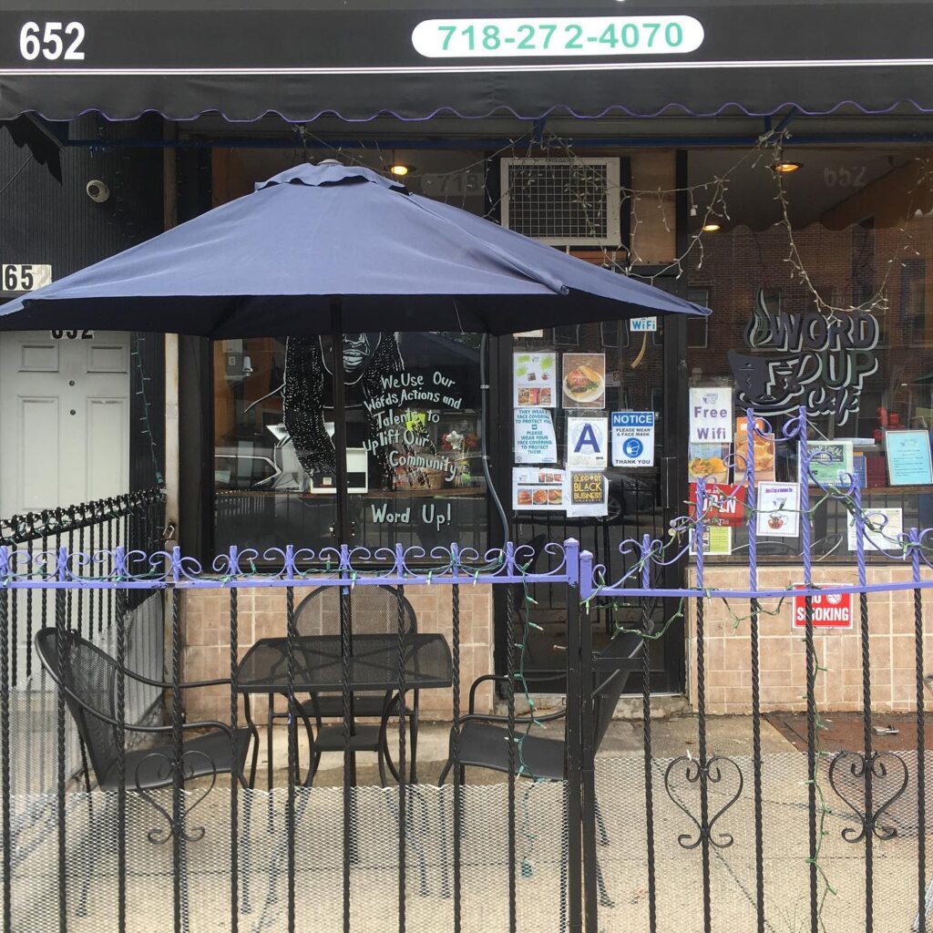 Restaurant exterior with glass windows and door; lawn umbrella, three chairs and a table all black enclosed by a black wrought iron fence at Word Up Cafea