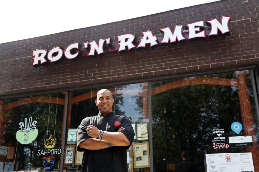 chef in a black tunic standing in front of a brick restaurant with glass windows at Roc N Ramen